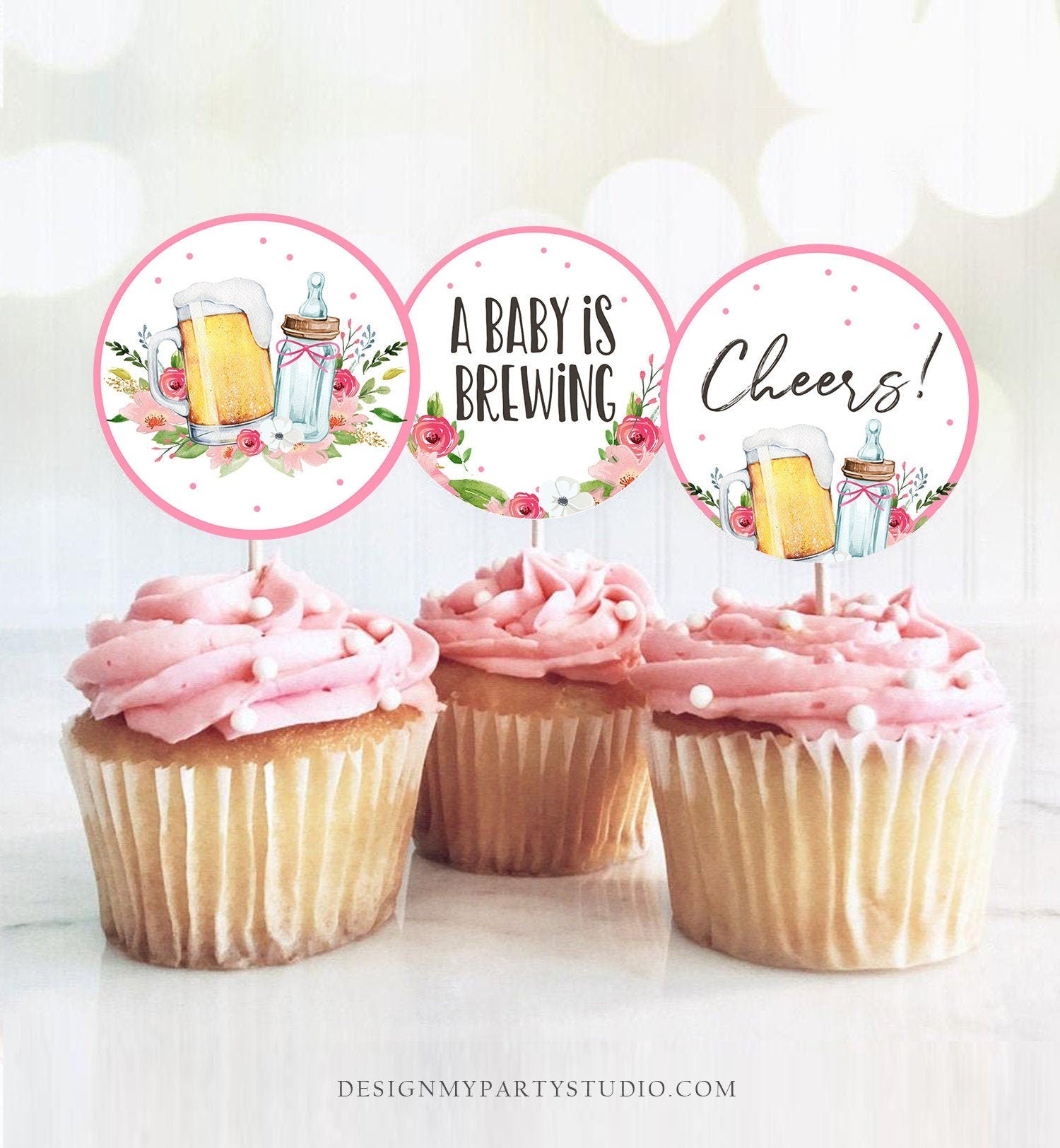 May include: Three pink and white cupcakes with round toppers. The toppers are white with pink borders and feature a beer mug, a mason jar, and floral designs. The text on the toppers reads 'A Baby is Brewing' and 'Cheers!'.
