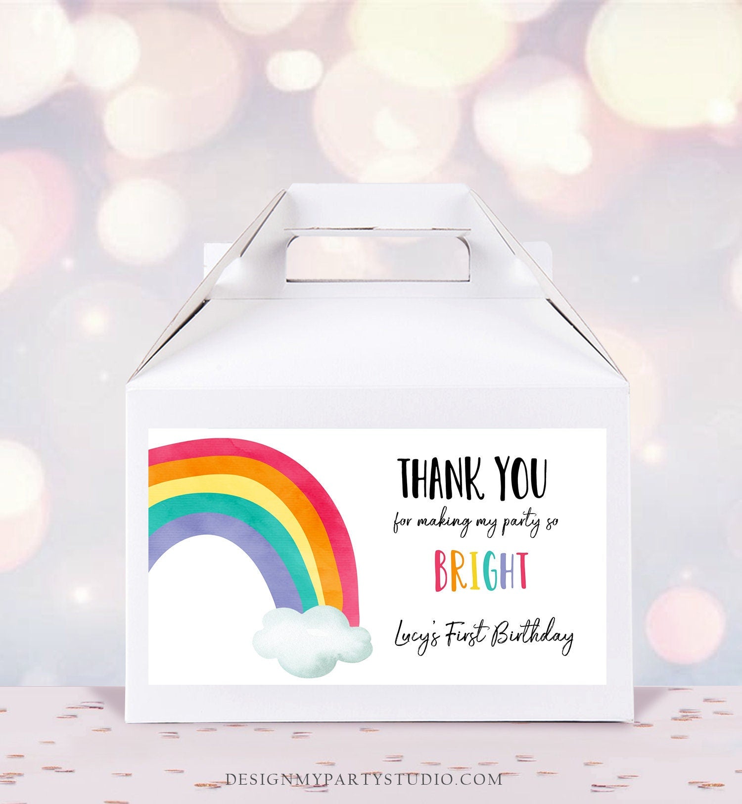 May include: A white favor box with a rainbow and cloud design and a thank you sticker that says 'Thank you for making my party so bright Lucy's First Birthday'.