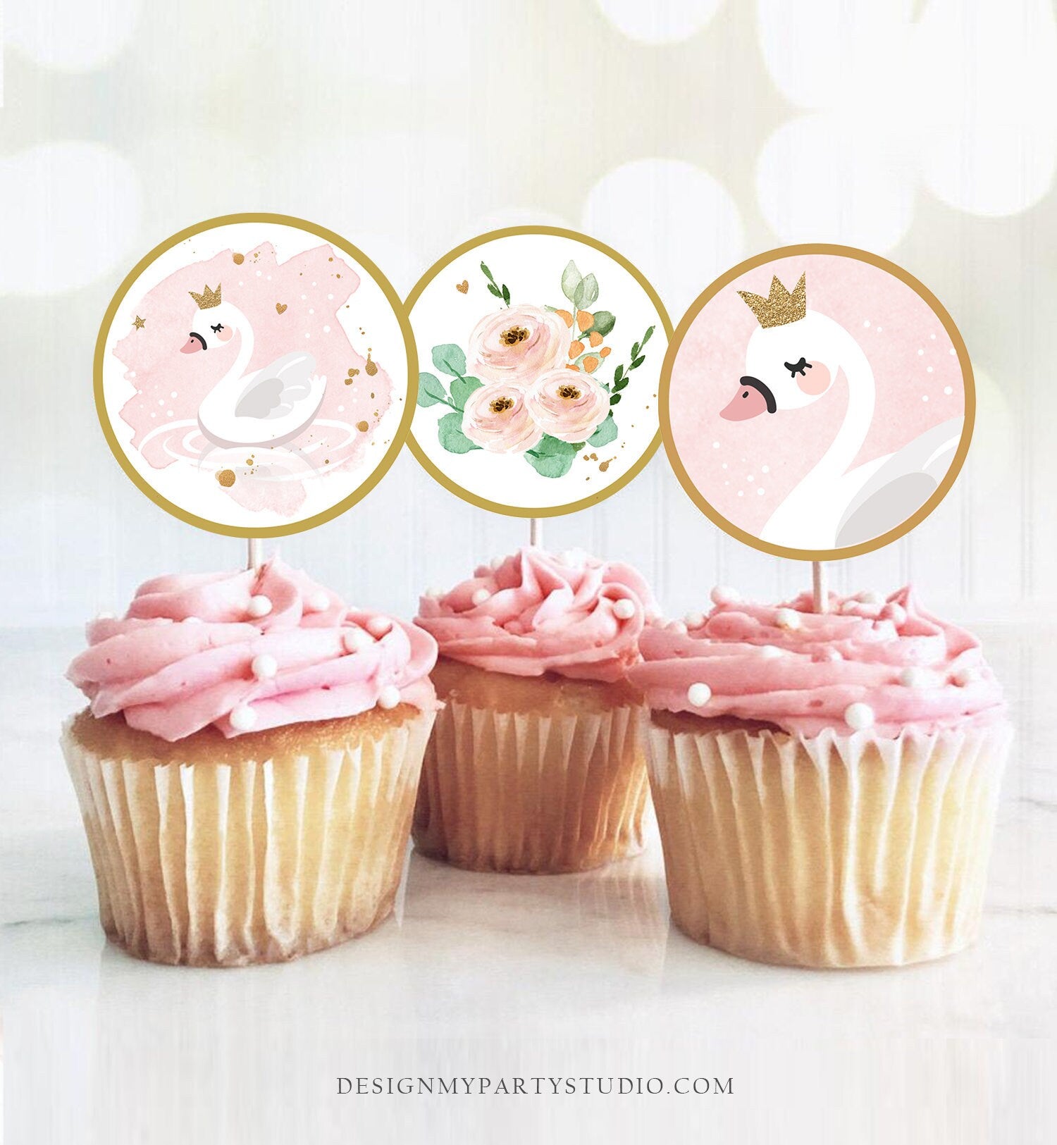 May include: Three pink and white cupcakes with white frosting and pearl sprinkles. Each cupcake has a round, gold-rimmed topper with a pink and white design. The toppers feature a white swan with a gold crown, a bouquet of pink and white flowers, and a white swan with a gold crown.