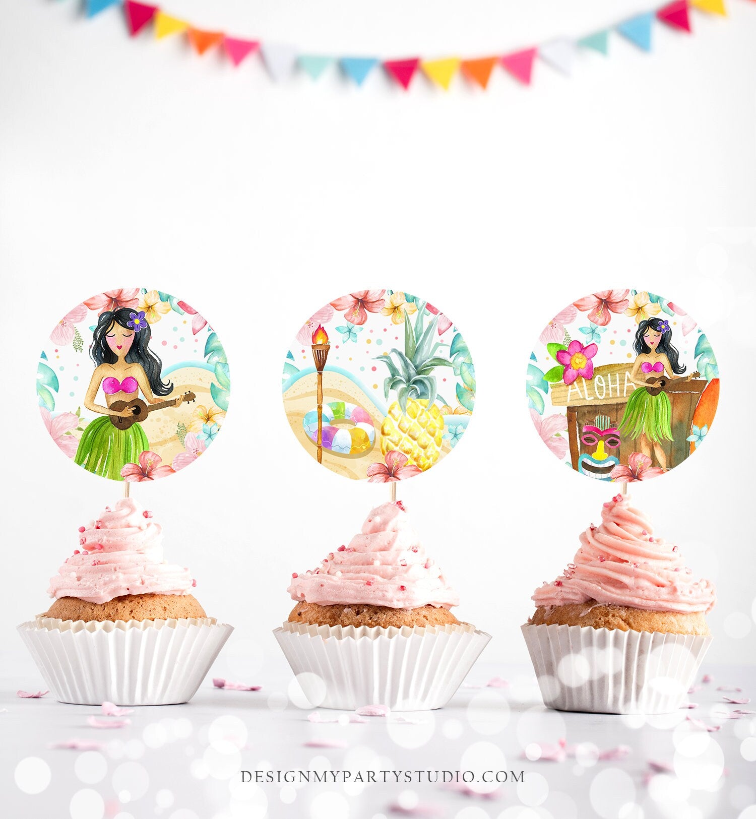 May include: Three cupcake toppers with a tropical theme. The toppers are round and feature a watercolor illustration of a woman in a hula skirt playing a ukulele, a pineapple, and a tiki hut with the word &#39;Aloha&#39; on it. The toppers are on top of cupcakes with different colored frosting.