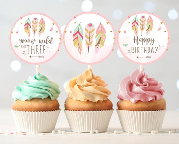 May include: Three pink and gold cupcake toppers with feathers and text. The toppers read &amp;#39;young and wild three&amp;#39;, &amp;#39;happy birthday&amp;#39;, and &amp;#39;happy birthday&amp;#39;. The toppers are on top of cupcakes with green, yellow, and pink frosting.