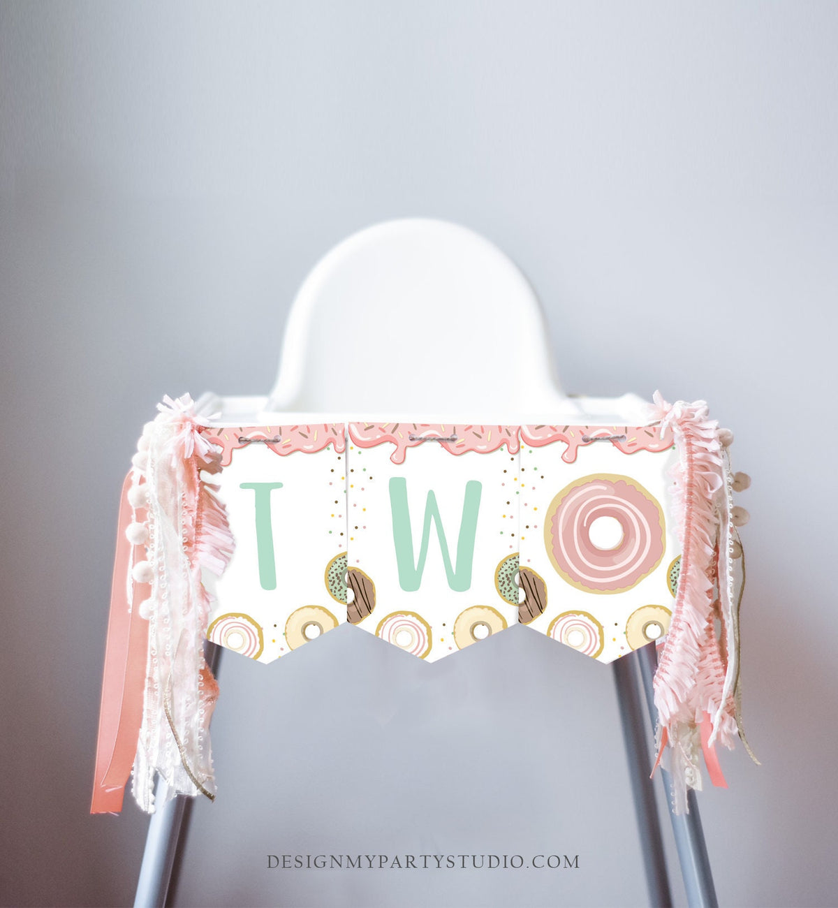 May include: A high chair with a pink and white donut themed banner that says &amp;#39;TWO&amp;#39; with sprinkles and a donut graphic. The banner has a scalloped edge and is decorated with pink and white ribbon and a white tassel garland.
