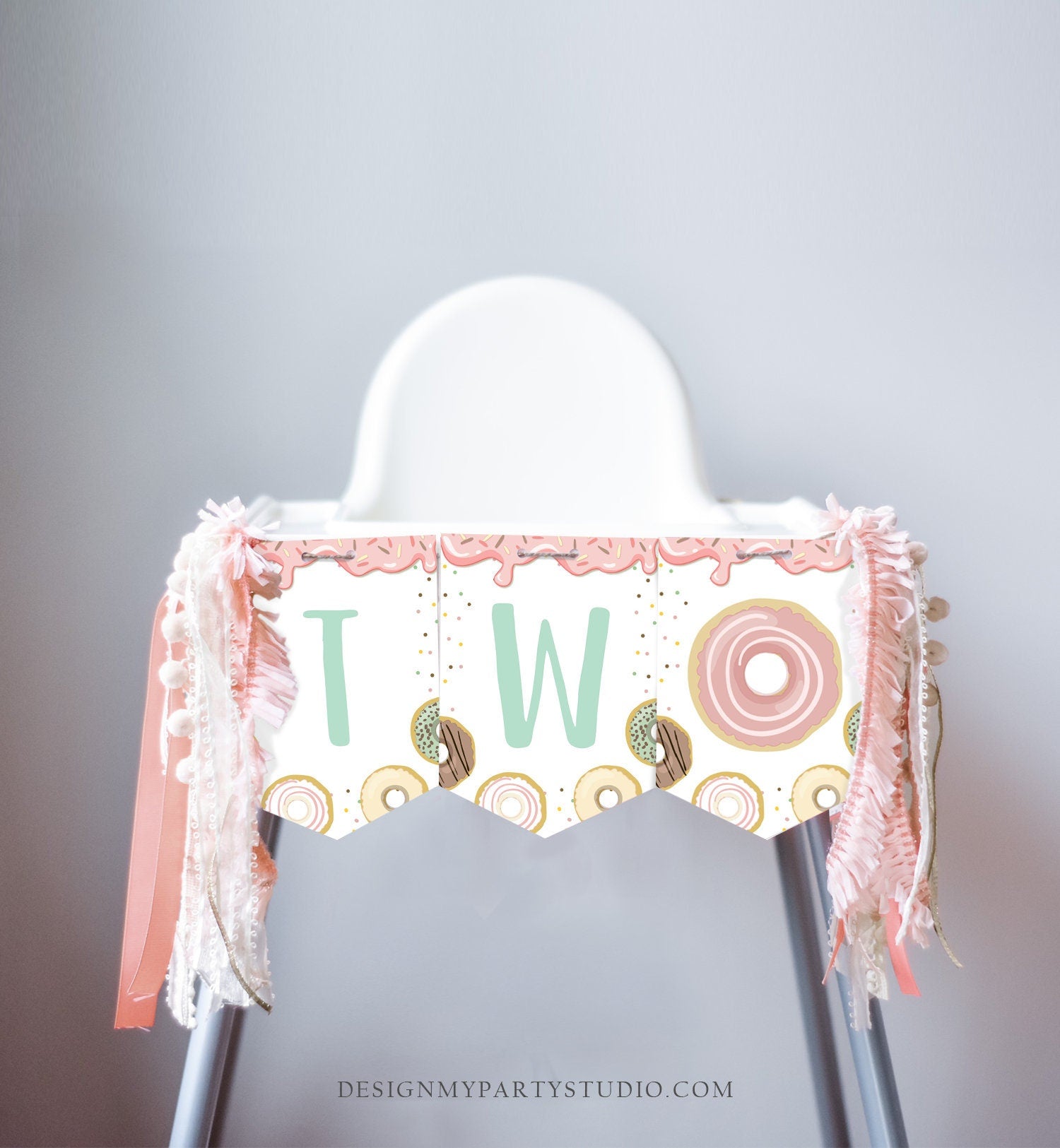 May include: A high chair with a pink and white donut themed banner that says &#39;TWO&#39; with sprinkles and a donut graphic. The banner has a scalloped edge and is decorated with pink and white ribbon and a white tassel garland.