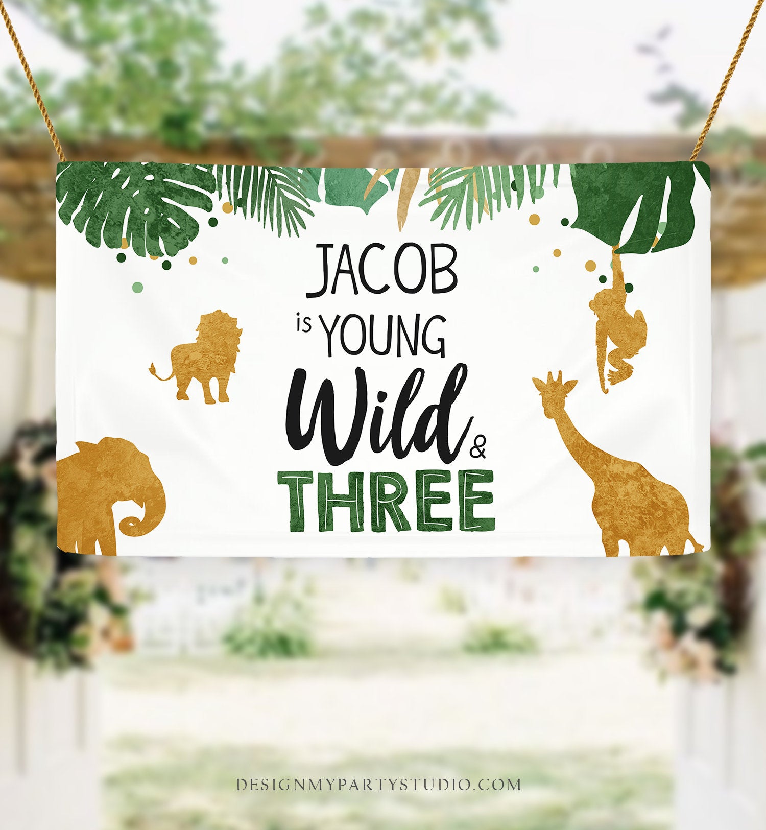 May include: A white banner with green and gold jungle animal silhouettes. The banner reads &#39;Jacob is young wild & three&#39; with the words &#39;wild&#39; and &#39;three&#39; in green and the rest in black. The banner is decorated with green leaves and gold confetti.