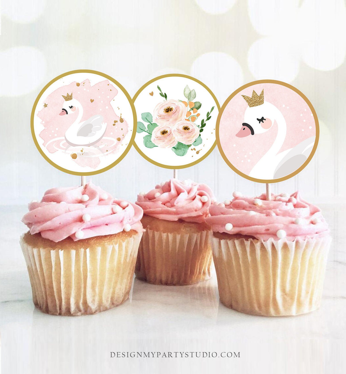 May include: Three pink and white cupcakes with white frosting and pearl sprinkles. Each cupcake has a round, gold-rimmed topper with a pink and white design. The toppers feature a white swan with a gold crown, a bouquet of pink and white flowers, and a white swan with a gold crown.